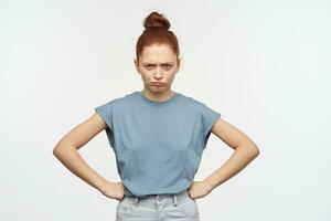 Portrait of upset, adult redhead girl with hair gathered in a bun. Wearing blue t-shirt and jeans. Put hands on her hips and frowning. Watching at the camera isolated over white background photo