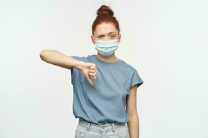 Portrait of disappointed redhead girl with hair gathered in a bun. Wearing blue t-shirt and protective mask. Showing thumb down in disagreement. Watching at the camera isolated over white background photo