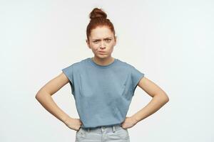 Serious looking woman, girl with ginger hair gathered in a bun. Wearing blue t-shirt and jeans. Put hand on her hips and frowns. Emotion concept. Watching at the camera isolated over white background photo