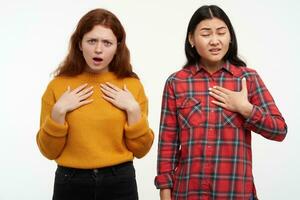 Two young, unhappy women friends. Confused, pointing doubted on themselves. People concept. Wearing yellow sweater and checkered shirt. Watching at the camera, isolated over white background photo