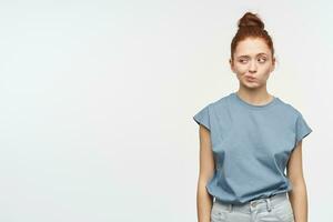 Nice looking woman, beautiful girl with ginger hair gathered in a bun. Wearing blue t-shirt and jeans. Squint her face and watching to the left at copy space, isolated over white background photo