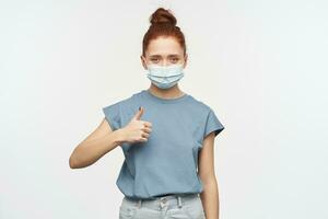 Nice looking woman, happy girl with ginger hair gathered in a bun. Wearing blue t-shirt, jeans and protective face mask. Showing thumb up. Watching at the camera isolated over white background photo