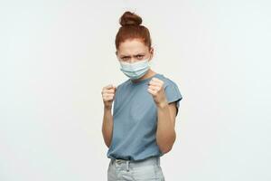 Portrait of angry redhead girl with hair gathered in a bun. Wearing blue t-shirt and protective face mask. Clench her fists, ready to fight. Watching at the camera isolated over white background photo