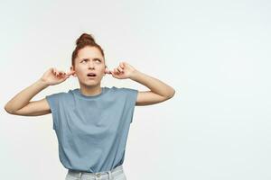 Unhappy looking woman with ginger hair gathered in a bun. Wearing blue t-shirt and jeans. Close her ears with fingers. Watching to the right upper corner at copy space, isolated over white background photo