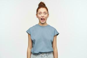 Shocked looking woman, beautiful girl with ginger hair gathered in a bun. Wearing blue t-shirt and jeans. Lick her lip, shows tongue. Watching at the camera isolated over white background photo