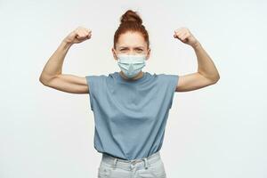 Teenage girl, strong woman with ginger hair gathered in a bun. Wearing protective face mask. Clench and raise her fists. Showing strength. Watching at the camera isolated over white background photo