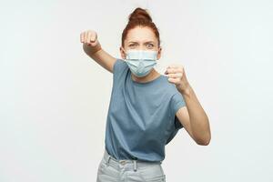 Aggressive looking woman with ginger hair gathered in a bun. Wearing blue t-shirt and protective face mask. Clench her fists and want to punch. Watching at the camera isolated over white background photo