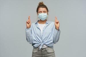 Happy, teenage girl with blond hair gathered in bun. Wearing striped knotted shirt and protective face mask. Keeps eyes closed, fingers crossed. Making a wish. Stand isolated over grey background photo