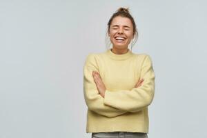 Portrait of cheerful, adult girl with blond hair gathered in bun. Wearing yellow sweater. Laughing with eyes closed and arms crossed on a chest. Stand isolated over white background photo