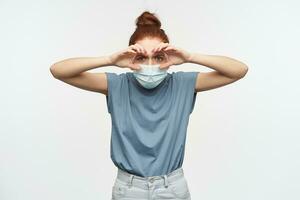 Nice looking woman, beautiful girl with ginger hair gathered in a bun. Wearing blue t-shirt, jeans and face mask. Watching at the camera isolated over white background photo