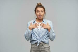 Teenage girl, excited looking woman with blond hair gathered in bun. Wearing striped knotted shirt. Puts palms on a chest, pointing at herself. Watching at the camera, isolated over grey background photo