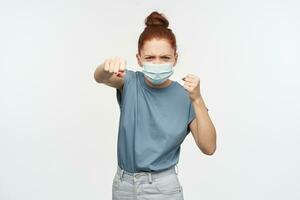 Angry looking woman, girl with ginger hair gathered in a bun. Wearing blue t-shirt, jeans and protective face mask. Clench her fists and punching. Watching at the camera isolated over white background photo