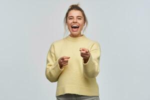 Cheerful woman, beautiful girl with blond hair gathered in bun. Wearing yellow sweater. Laughing from you. Pointing with fingers and watching at the camera, isolated over white background photo