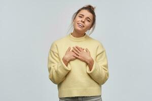 Teenage girl, charming woman with blond hair gathered in bun. Wearing yellow sweater. Tilt her head and puts palms on her chest. Hear compliment. Watching at the camera, isolated over white background photo