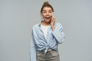 Excited, happy looking woman with blond hair gathered in bun. Wearing striped knotted shirt and talking on her cellphone. Hear great news. Watching at the camera, isolated over grey background photo