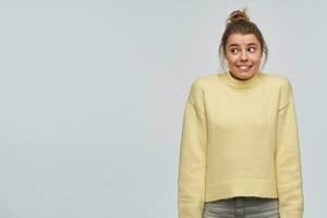 Nice looking woman, beautiful girl with blond hair gathered in bun. Wearing yellow sweater. Shrugs her shoulders and biting a lip. Watching to the left at copy space, isolated over white background photo