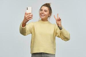 Portrait of attractive, adult girl with blond hair gathered in bun and tattoo. Wearing yellow sweater and holding a smartphone. Making selfie. Showing peace sign. Stand isolated over white background photo