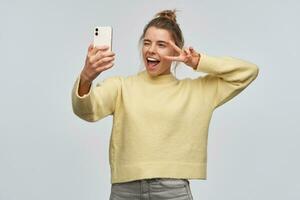 Cheerful looking woman with blond hair gathered in bun. Wearing yellow sweater and holding a smartphone. Making selfie. Showing peace sign over her eye and winks. Stand isolated over white background photo