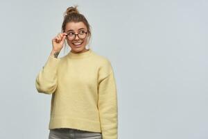 Portrait of attractive, cute girl with blond hair gathered in bun. Wearing yellow jumper and eyewear. Touching her glasses and gaze over them to the right at copy space, isolated over white background photo