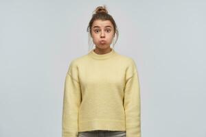 Portrait of attractive, funny girl with blond hair gathered in bun. Wearing yellow sweater. Pouts her cheeks. Emotion concept. Watching at the camera, isolated over white background photo