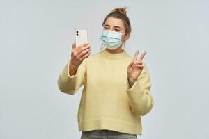 Positive, beautiful girl with blond hair gathered in bun. Wearing yellow sweater and protective face mask. Making selfie on a smartphone and showing peace sign. Stand isolated over white background photo