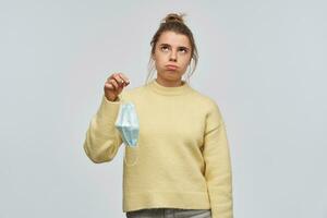 Annoyed girl with blond hair gathered in bun. Wearing yellow sweater. Holding protective face mask. Breathe out, exhausted from wearing mask. Watching up at copy space, isolated over white background photo