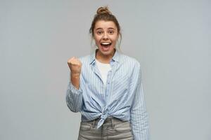 Portrait of attractive, excited girl with blond hair gathered in bun. Wearing striped knotted shirt. Clench her fist and celebrate success. Watching at the camera, isolated over grey background photo