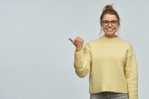 Smiling, beautiful girl with blond hair gathered in bun. Wearing yellow sweater and eyewear. Pointing with thumb to the left at copy space. Watching at the camera, isolated over white background photo