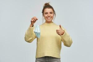 Teenage girl, happy looking woman with blond hair gathered in bun. Wearing yellow sweater. Holding protective face mask and showing thumb up. Watching at the camera, isolated over white background photo