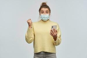 Excited, happy woman with blond hair gathered in bun. Wearing yellow sweater and protective face mask. Holding a smartphone and clench her fist. Watching at the camera, isolated over white background photo