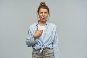 Unhappy woman, angry girl with blond hair gathered in bun. Wearing striped knotted shirt. Frowns in confusion and pointing at herself. Watching at the camera, isolated over grey background photo