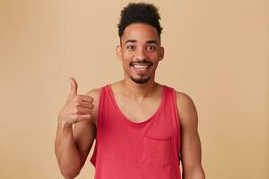 Portrait of handsome, happy african american male with afro hairstyle and beard. Wearing red tank-top. Showing thumb up sign, all good. Watching at the camera isolated over pastel beige background photo