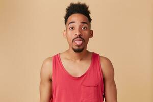 Teenage african american, funny looking man with afro hairstyle and beard. Wearing red tank-top. Showing tongue, playful mood. Watching at the camera isolated over pastel beige background photo