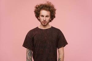 Cool looking man, handsome guy with tattoos, brunette curly hair and beard. Wearing dark red t-shirt. Emotion and people concept. Watching serious at the camera isolated over pastel pink background photo