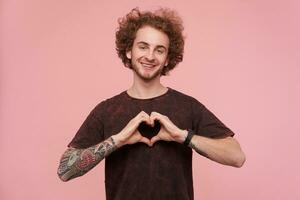 Young, happy man with brunette curly hair, beard and tattoos. Wearing dark red t-shirt. Emotion concept. Showing love sign with fingers. Watching at the camera isolated over pastel pink background photo