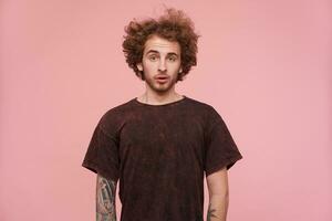 Portrait of handsome man with brunette curly hair, beard and tattoos. Wearing dark red t-shirt. Emotion concept. Looking slightly surprised. Watching at the camera isolated over pastel pink background photo