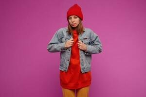 Teenage girl, suspicious looking woman with brunette long hair. Wearing jeans jacket, yellow pants, red sweater and hat. Straighten jacket. Watching at the camera isolated over purple background photo