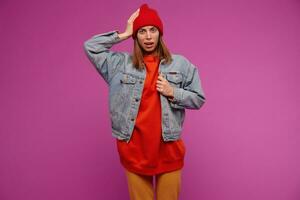Bewildered looking woman with brunette hair. Wearing jeans jacket, yellow pants, red sweater and hat. Touching her head, disconnected. Watching at the camera isolated over purple background photo