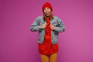 Funny looking woman with brunette hair. Wearing jeans jacket, yellow pants, red sweater and hat. Shrugs, straighten the jacket, show tongue. Watching up at copy space, isolated over purple background photo