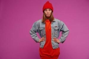 Funny looking woman, playful girl with brunette hair. Wearing jeans jacket, red sweater and hat. Put hands on a waist a showing tongue. Watching at the camera isolated over purple background photo