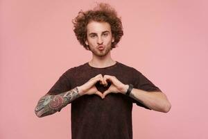 Handsome guy with tattoos, brunette curly hair and beard. Wearing dark red t-shirt. Showing love sign with fingers and try to kiss. Watching at the camera isolated over pastel pink background photo