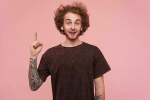 Young man, cool guy with brunette curly hair, beard and tattoos. Wearing dark red t-shirt. Emotion concept. Rise finger up, got an idea. Watching at the camera isolated over pastel pink background photo