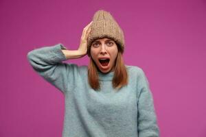 Portrait of horrified, young woman with brunette long hair. Wearing blue sweater and knitted hat. Emotion concept. Touching her head and screams. Watching at the camera isolated over purple background photo