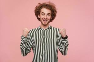 Young man, excited guy with brunette curly hair and beard. Wearing shirt with black and white stripes and watch. Clinch hes fists. Watching happy at the camera isolated over pastel pink background photo