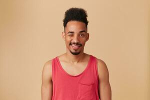 Portrait of handsome, african american adult male with afro hairstyle and beard. Wearing red tank-top. Emotion concept. Watching and wink at the camera isolated over pastel beige background photo