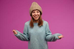 Teenage girl, happy looking woman with brunette long hair. Wearing blue sweater and knitted hat. Emotion concept. Shrugs her hands in disbelief. Watching at the camera isolated over purple background photo