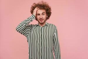 Portrait of tired, young man with brunette curly hair and beard. Wearing shirt with black and white stripes. Touching hes head. Forget. Watching at the camera isolated over pastel pink background photo