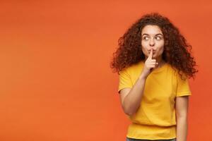 Portrait of attractive, redhead girl with curly hair. Wearing yellow t-shirt. People and emotion concept. Showing silence sign. Watching to the left at copy space, isolated over orange background photo