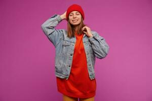 Charming looking woman, beautiful girl with brunette hair. Wearing jeans jacket, yellow pants, red sweater and hat. Touching her head and keep eyes closed. Stand isolated over purple background photo