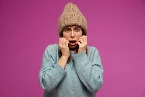 Afraid looking woman, terrified girl with brunette hair. Wearing blue sweater and knitted hat. Emotion concept. Touching her face in fear. Watching at the camera isolated over purple background photo
